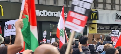 Crowd of people holding various protest signs and flags in front of McDonald's and Itsu restaurants. Some signs have messages related to stopping a massacre and proclaiming freedom for Palestine.