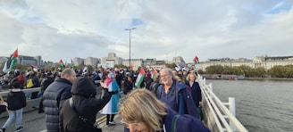 A large group of people are gathered on a bridge, many holding flags with red, white, green, and black colors. The atmosphere appears lively and crowded, with buildings visible in the background along a riverside. The sky is cloudy, and everyone is dressed warmly, suggesting cool weather.
