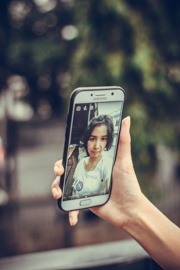 A hand holds a smartphone displaying a selfie on its screen. The selfie captures a person wearing a white shirt with a badge, in an indoor environment with blurred background details. The surrounding environment appears natural and green.