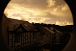 The image depicts a serene village scene with traditional houses featuring gabled roofs and lattice wall patterns. The sky is filled with dramatic clouds, taking on a golden hue as the sun sets behind a silhouette of hills.