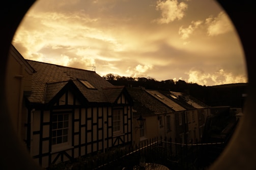 The image depicts a serene village scene with traditional houses featuring gabled roofs and lattice wall patterns. The sky is filled with dramatic clouds, taking on a golden hue as the sun sets behind a silhouette of hills.