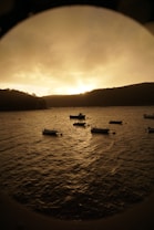 The image captures a serene waterfront view during sunset. Small boats drift gently on a rippling body of water. The sky is filled with a warm, golden hue, creating a peaceful and tranquil atmosphere. Hills in the background are silhouetted against the fading light.