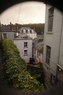 A quaint, old town alleyway lined with ivy-covered walls and brick buildings. The buildings have multiple stories, some with dormer windows, and appear historic with classic architectural elements. The alley is narrow and there is a glimpse of greenery in the background with a slight view of distant hills.