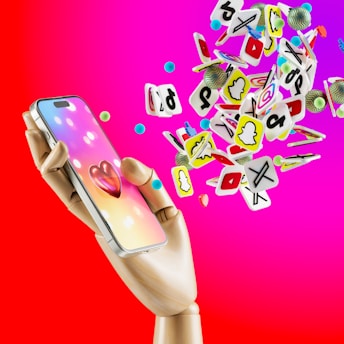 A wooden mannequin hand is holding a smartphone with a colorful screen displaying a 3D heart icon. Surrounding the phone are numerous floating social media icons in various shapes and colors, set against a vibrant pink and red gradient background.