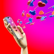 A wooden mannequin hand holding a smartphone, with heart and like icons emerging from the screen against a bright pink and red background. The icons appear three-dimensional and colorful, symbolizing social media engagement.