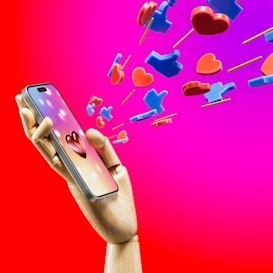 A wooden mannequin hand holding a smartphone, with heart and like icons emerging from the screen against a bright pink and red background. The icons appear three-dimensional and colorful, symbolizing social media engagement.