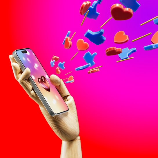 A wooden mannequin hand holding a smartphone, with heart and like icons emerging from the screen against a bright pink and red background. The icons appear three-dimensional and colorful, symbolizing social media engagement.