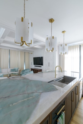 A modern kitchen area with a large island featuring a sink and gold faucet. The countertop has a smooth, stone-like slab with light green and beige tones. Above the island, four white pendant lights with gold accents hang from the ceiling. The background includes a living area with a cream sofa, teal cushions, and a mounted flat-screen TV. Large windows with sheer curtains allow natural light to flood the space.
