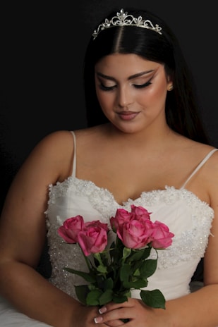 A woman with long dark hair is adorned with an elegant tiara and wearing a beaded white gown. She is holding a bouquet of vibrant pink roses and looking down softly.