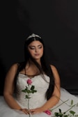 A woman wearing a white, beaded gown with a tiara in her long black hair. She is holding a pink rose and sitting against a dark background. Her eyes are closed and her expression is serene.
