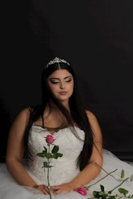 A woman wearing a white, beaded gown with a tiara in her long black hair. She is holding a pink rose and sitting against a dark background. Her eyes are closed and her expression is serene.