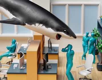 In an office setting, a large shark floats above workstations, surrounded by blue humanoid figures. One figure appears to be startled with hands on their head, while others stand nearby. The office has light wood flooring, yellow partitions, white chairs, and computer desks. Plants are visible in the background.