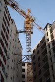 A tall yellow construction crane is positioned between two high-rise buildings. The buildings appear to be under construction, with some windows installed and others open. The scene is dominated by the urban architecture and industrial machinery.