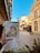 A person holds a small painting or sketch depicting a historic building with a dome and ornate details. The artwork is set against a real-life background of a cobblestone street flanked by old architectural structures. Trees and a clear blue sky add to the scene, creating a harmonious blend between the art and reality. Shadows suggest a bright, sunny day, enhancing the historic and artistic atmosphere.