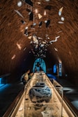 An intricately designed indoor exhibit featuring a long, glass-covered display table filled with various ceramic artifacts. Above the display, numerous fragmented pieces of ceramics hang from the high, arched brick ceiling. Several people are observing the artifacts, while the background is illuminated with ambient blue light from a large window and a digital display.