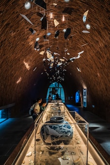 An intricately designed indoor exhibit featuring a long, glass-covered display table filled with various ceramic artifacts. Above the display, numerous fragmented pieces of ceramics hang from the high, arched brick ceiling. Several people are observing the artifacts, while the background is illuminated with ambient blue light from a large window and a digital display.