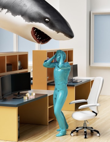 A surreal office scene featuring a large shark seemingly emerging from one of the walls above a workstation. A humanoid figure with a shiny teal appearance stands in a pose of shock or surprise. The office includes desks with computer monitors, a white office chair, and shelves. There are blocks on one desk spelling out 'FRED'.