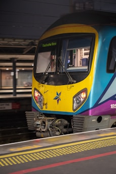 A passenger train is stationary at a platform, featuring a distinctive yellow and blue front with a purple accent and a decorative emblem. The electronic display indicates a destination of Manchester Airport. The platform has a visible yellow safety line and dark surroundings suggest it might be evening or night.