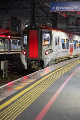 A modern train is stationed at a railway platform at night. The train features a sleek design with a prominent red and white color scheme. Overhead, the geometric structure of the station is visible, and a sign indicates 'Platform 13'. The platform is marked with safety lines for passengers.