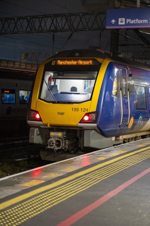 A modern passenger train is stationary on a platform, with a display indicating its destination as Manchester Airport. The train is primarily blue and yellow, with lighting that suggests it is evening or nighttime. Overhead wires are visible, suggesting an electric railway system.