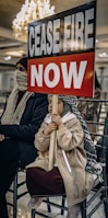 A child is sitting on a chair holding a sign that reads 'CEASE FIRE NOW'. The child is dressed warmly, with a headscarf and coat. There is an adult beside the child, also dressed warmly, partially covering their face. The background is indoors with a chandelier and row of chairs.