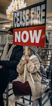 A child is sitting on a chair holding a sign that reads 'CEASE FIRE NOW'. The child is dressed warmly, with a headscarf and coat. There is an adult beside the child, also dressed warmly, partially covering their face. The background is indoors with a chandelier and row of chairs.
