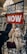 A child is sitting on a chair holding a sign that reads 'CEASE FIRE NOW'. The child is dressed warmly, with a headscarf and coat. There is an adult beside the child, also dressed warmly, partially covering their face. The background is indoors with a chandelier and row of chairs.