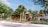 A sunny outdoor scene featuring a wooden pergola structure surrounded by palm trees and other lush greenery. Several people are walking and interacting in a landscaped area with paved walkways and decorative plants.
