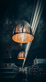 A dimly lit supermarket aisle with intricate pendant lights hanging from the ceiling. The lights have decorative cutouts that cast patterns on the surroundings. The shelves are stocked with various products, and the dark atmosphere is emphasized by limited illumination and shadows.