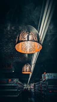 A dimly lit supermarket aisle with intricate pendant lights hanging from the ceiling. The lights have decorative cutouts that cast patterns on the surroundings. The shelves are stocked with various products, and the dark atmosphere is emphasized by limited illumination and shadows.