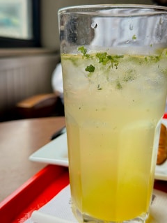 A tall glass filled with a refreshing lemon-lime beverage, garnished with mint leaves, is placed on a red tray. The tray rests on a wooden table next to a white plate. Sunlight filters through a nearby window, casting a soft glow on the drink.