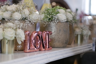 A collection of white roses and small white flowers arranged in rustic vases on a shelf. In the foreground, metallic rose gold letters spell the word LOVE. The setting is elegant and serene, suggesting a decorative arrangement for a special occasion.