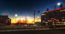A cityscape at dusk with a colorful sunset sky in the background. Urban buildings are illuminated with artificial lights, and a large building with signage is visible. Trees and streetlights line the streets, with traffic lights and cars present.