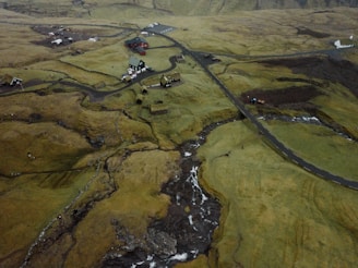 An aerial view of a rural landscape with a collection of scattered houses and buildings, surrounded by vast fields and winding roads. The terrain appears uneven with patches of grass and areas of exposed earth. A small stream runs through the landscape, with several bridges visible. The overall scene is set against a backdrop of hilly terrain.