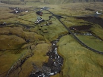 An aerial view of a rural landscape with a collection of scattered houses and buildings, surrounded by vast fields and winding roads. The terrain appears uneven with patches of grass and areas of exposed earth. A small stream runs through the landscape, with several bridges visible. The overall scene is set against a backdrop of hilly terrain.