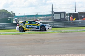 A race car with the number 777 is speeding along a racetrack. The car is covered in various advertisements and logos, including 'CarStore' prominently displayed on the side. The racetrack is bordered by green grass and a metal guardrail, and there are a few people in the background wearing orange safety suits.
