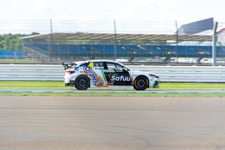 A race car with various sponsor logos is speeding along a track. The background includes blurred fencing and bleachers, indicating motion and speed.