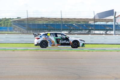 A race car with various sponsor logos is speeding along a track. The background includes blurred fencing and bleachers, indicating motion and speed.