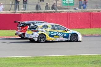Two race cars are competing side by side on a track. One car is predominantly blue and white with various sponsor logos, and the other is red with black and white accents. The background has a red barrier and a fence with spectators visible behind it.