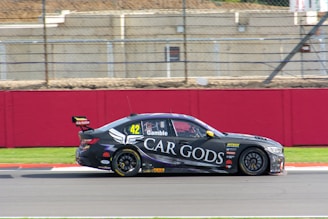 A sleek black race car speeds along the track with sponsorship branding prominently displayed on its side. The car is numbered 42 and features logos and text in white and yellow accents. It races past a red barrier with a wire fence and buildings in the background.