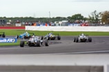 Several race cars navigate a track during a motorsport event. The cars are captured making a turn on the asphalt with a clear view of the rear wings and tire details. Spectators are visible in the background, some standing along the barriers, while others sit on the viewing stands. The scene is lively with bright colors and racing decals on the cars.
