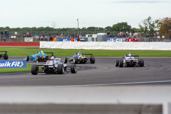 Several race cars navigate a track during a motorsport event. The cars are captured making a turn on the asphalt with a clear view of the rear wings and tire details. Spectators are visible in the background, some standing along the barriers, while others sit on the viewing stands. The scene is lively with bright colors and racing decals on the cars.