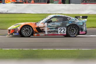 A colorful racing car, number 22, is speeding on a race track. The car's exterior features a vibrant and artistic design with a combination of geometric patterns and various logos. It has a large rear wing and sleek aerodynamic body, set against a background that includes grass and a barrier with spectators behind.