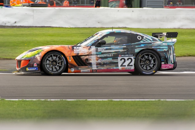 A colorful racing car, number 22, is speeding on a race track. The car's exterior features a vibrant and artistic design with a combination of geometric patterns and various logos. It has a large rear wing and sleek aerodynamic body, set against a background that includes grass and a barrier with spectators behind.