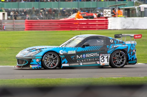 A blue race car with various sponsor logos and the number 31 is driving on a racetrack. The car is adorned with patterns and decals, including a large rear spoiler. In the background, there is a red barrier, green grass, and a crowd of spectators. Additionally, there is a person in an orange safety vest near the barrier.