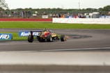 A race car navigating a curve on a motorsport track, surrounded by grassy areas, with spectators visible in the background behind a safety barrier. The car is painted in bright colors and is equipped with slick tires. Advertisement banners are positioned along the edge of the track.