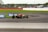 A race car navigating a curve on a motorsport track, surrounded by grassy areas, with spectators visible in the background behind a safety barrier. The car is painted in bright colors and is equipped with slick tires. Advertisement banners are positioned along the edge of the track.