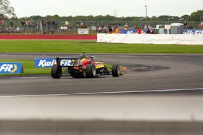 A race car navigating a curve on a motorsport track, surrounded by grassy areas, with spectators visible in the background behind a safety barrier. The car is painted in bright colors and is equipped with slick tires. Advertisement banners are positioned along the edge of the track.