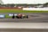 A race car navigating a curve on a motorsport track, surrounded by grassy areas, with spectators visible in the background behind a safety barrier. The car is painted in bright colors and is equipped with slick tires. Advertisement banners are positioned along the edge of the track.