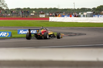 A race car navigating a curve on a motorsport track, surrounded by grassy areas, with spectators visible in the background behind a safety barrier. The car is painted in bright colors and is equipped with slick tires. Advertisement banners are positioned along the edge of the track.